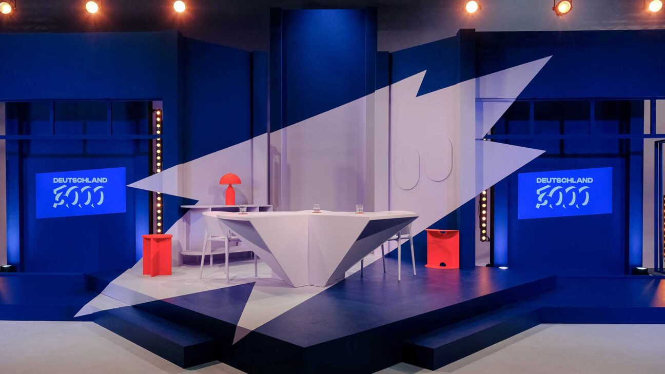 Modernes Fernsehstudio-Set mit geometrischem Design, blauen und weißen Elementen, einem dreieckigen Tisch, roten Akzenten wie einer Lampe und einem Hocker sowie dem 'Deutschland 3000'-Logo, das prominent auf Bildschirmen im Hintergrund angezeigt wird.